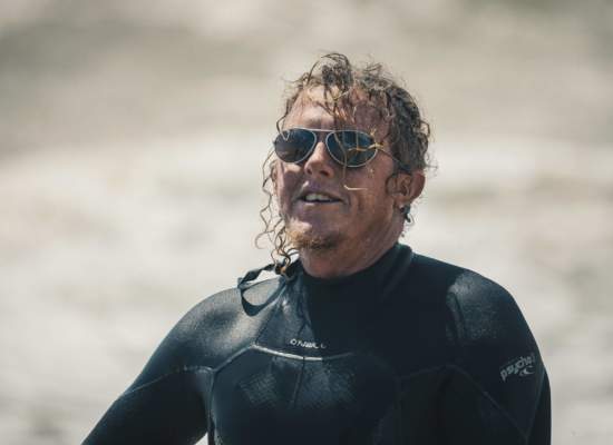 Man with long curly hair, coming out of the sea wearing a wetsuit and Bigatmo sunglasses.